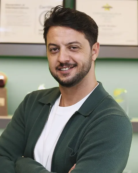 Olcay Silahlı, koyu yeşil ceket ve beyaz tişörtle, kolları kavuşturmuş gülümserken.